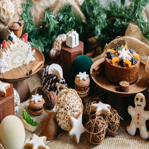 A table displays cakes, cookies, macarons, and seasonal treats on wooden stands with surrounding greenery and ornaments for a mindful holiday celebration.