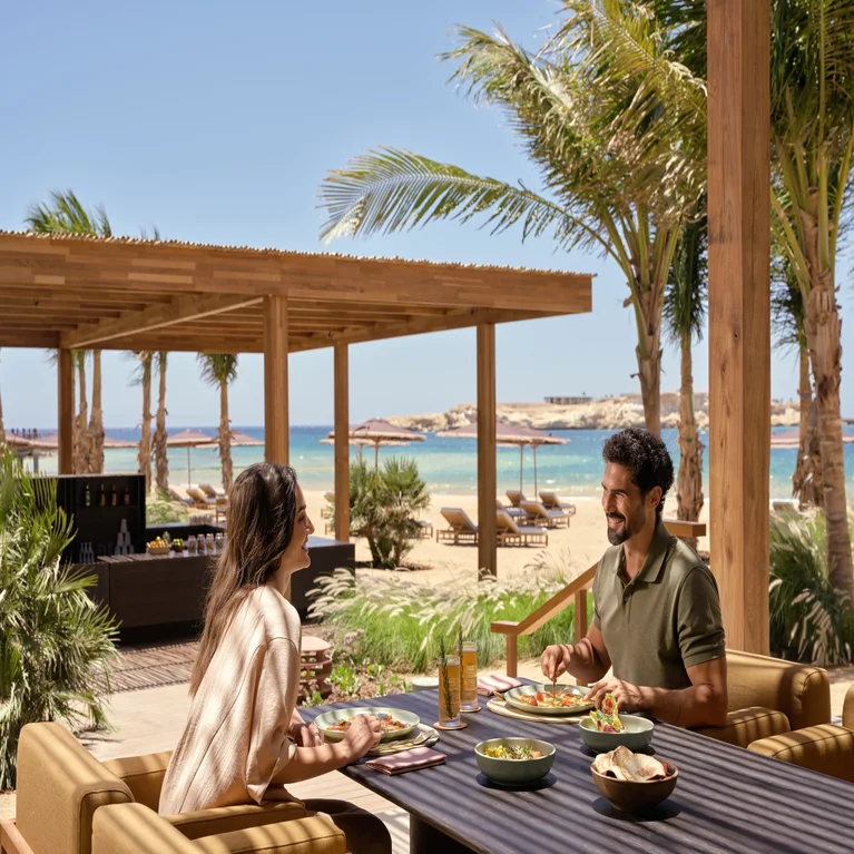 Guests share a meal at an open-air restaurant shaded by palm trees, with views of a sandy shore and the ocean beneath a clear sky.