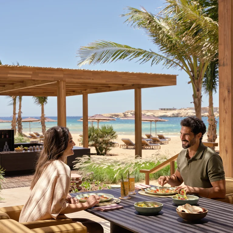Guests share a meal at an open-air restaurant shaded by palm trees, with views of a sandy shore and the ocean beneath a clear sky.