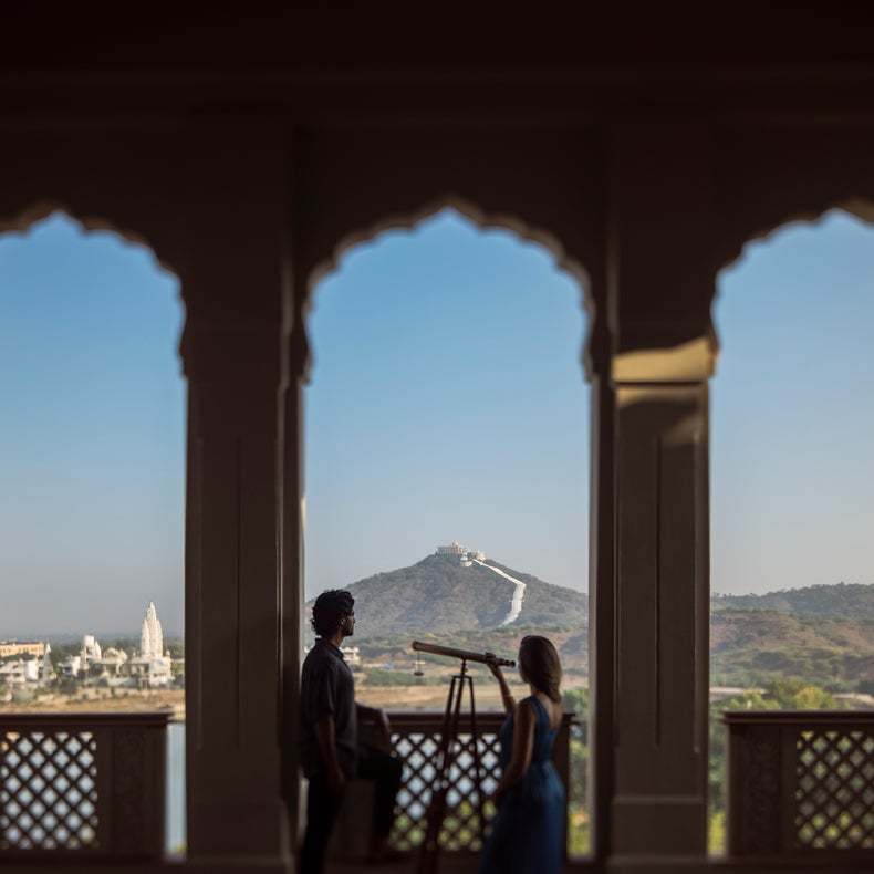 Los huéspedes observan un histórico fuerte en la cima de una colina y el paisaje urbano a través de un telescopio desde un balcón con ventana arqueada, con el cielo abierto de fondo.