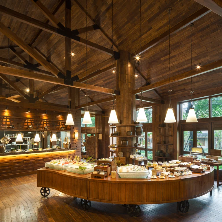 Un restaurante con vigas y suelos de madera natural, iluminación colgante, amplios ventanales y un bufé central con una selección de alimentos frescos.