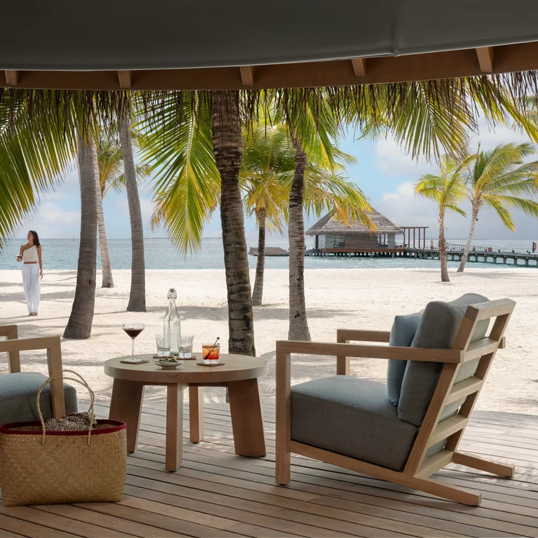 تم تأثيث الشرفة المظلّلة بكراسي بذراعين وطاولة مع مشروبات. يمكنك أن ترى من بعيد شاطئاً رملياً وأشجار نخيل ورصيفاً خشبياً ورجل يتنزه على طول الشاطئ.....