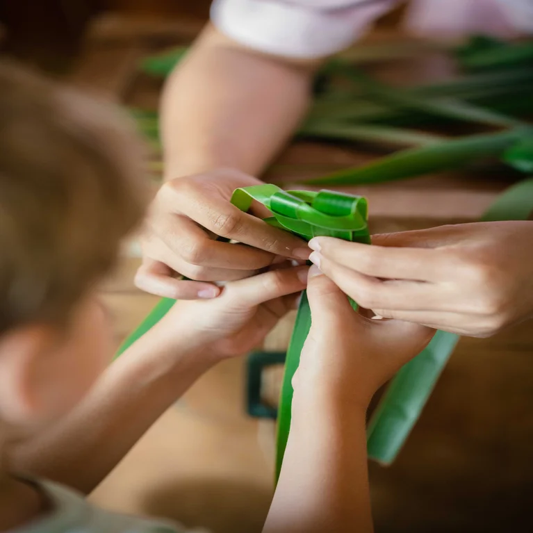 Adult hands gently guiding a child in weaving green palm leaves, reflecting shared learning and connection to nature.