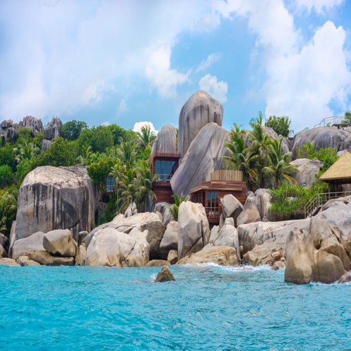A house built from natural materials sits among granite boulders and palms beside clear turquoise water, beneath a sky with scattered clouds.