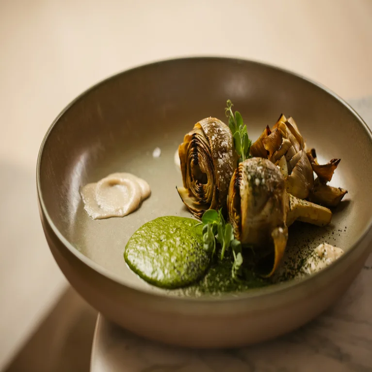 Two roasted artichokes with green and beige sauces, paired with fresh greens on a ceramic dish. A balanced presentation emphasizing wellness and natural ingredients.