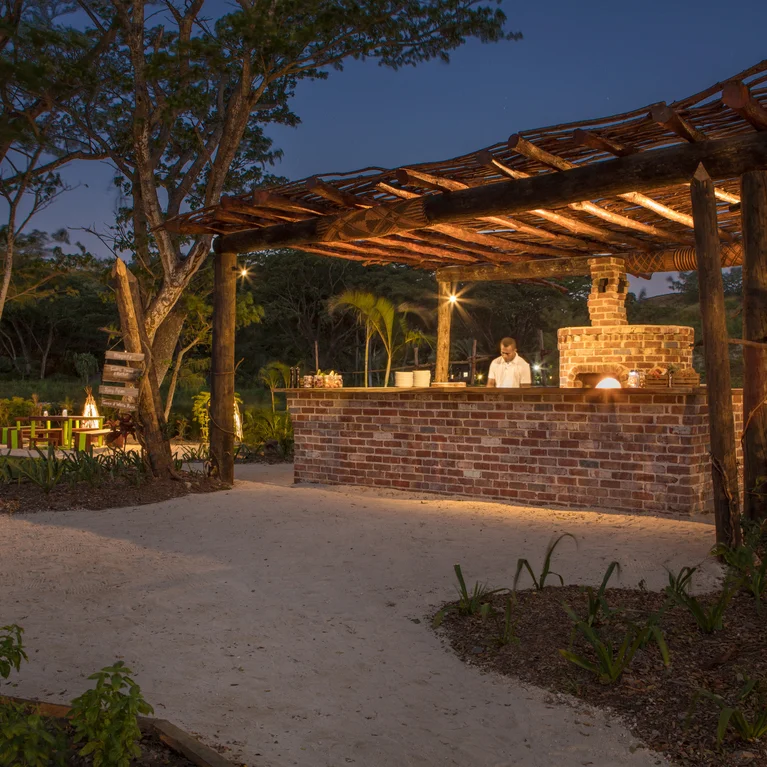 Sustainable bar under a wooden roof and brick counter, softly illuminated at dusk. Surrounded by greenery, team member present behind the counter.