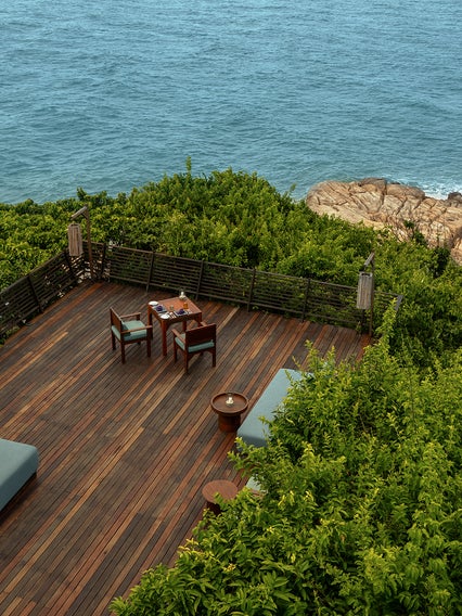 Deck with seating and lounge chairs faces the ocean, bordered by green bushes and natural rocky shore, offering a calm setting for relaxation and connection to nature.