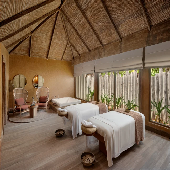 Spa room featuring two massage tables, a sunken tub, seating area, and large windows offering abundant natural light and views of surrounding plants.