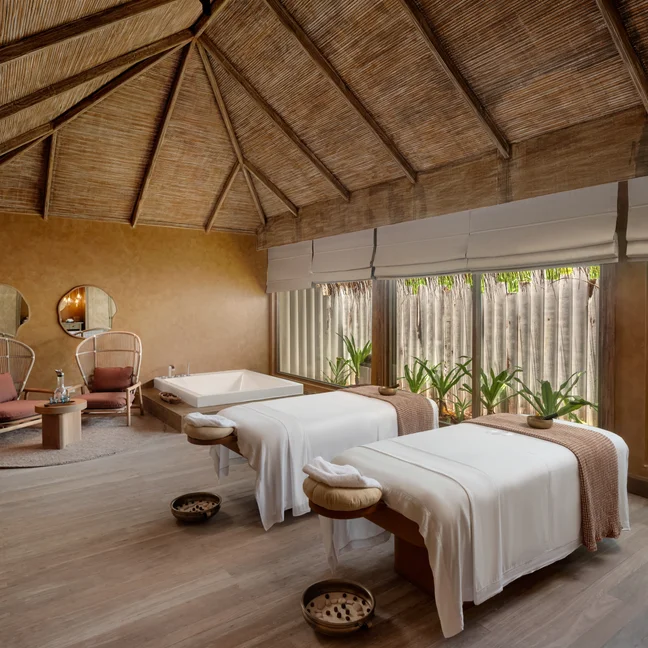 Spa room featuring two massage tables, a sunken tub, seating area, and large windows offering abundant natural light and views of surrounding plants.