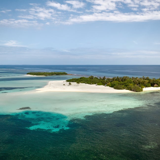 An island with soft sand, vibrant greenery, and clear blue water lies in the ocean beneath a sky spotted with clouds.