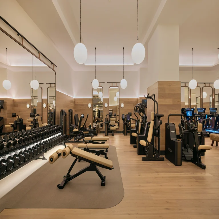 Wellness-focused fitness space featuring neutral-toned benches, free weights, equipment, wood flooring and ample lighting for a calm, accessible workout environment.