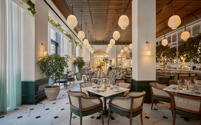 Restaurant space with marble floors, abundant greenery, circular pendant lights, and tables prepared with glassware and plates in an open, sunlit environment.