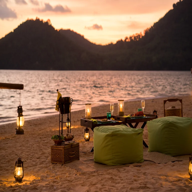 Sustainable picnic arrangement with green bean bags, a low table with local cuisine and drinks, lantern lighting, and mountain views at dusk.