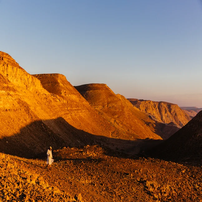 Individual in light clothing stands on sunlit rocky ground, surrounded by golden desert hills beneath a clear blue sky.