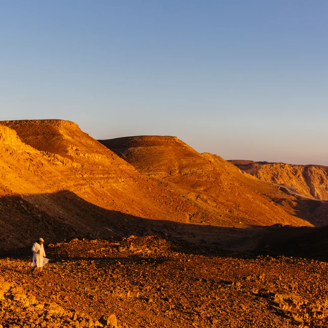 Individual in light clothing stands on sunlit rocky ground, surrounded by golden desert hills beneath a clear blue sky.