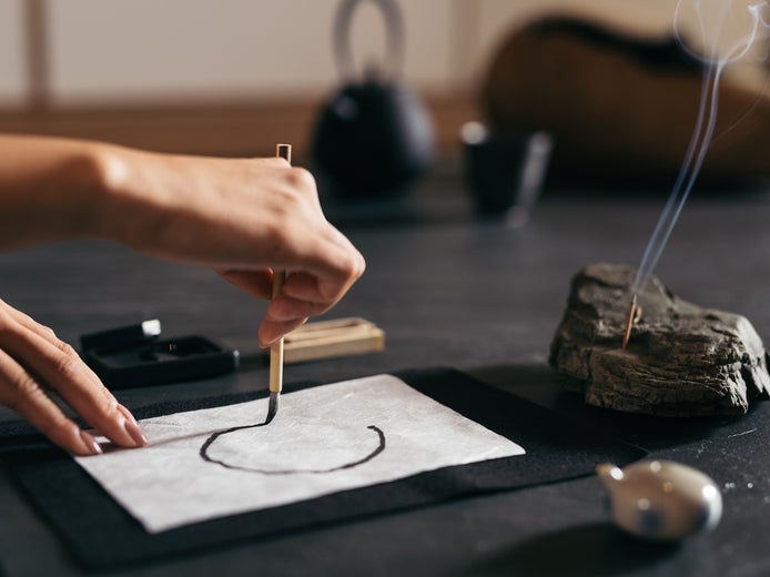 Một người vẽ một nét cọ tròn bằng mực đen trên giấy trắng, với nhang và các dụng cụ nghệ thuật truyền thống được sắp xếp trên bàn để tạo ra một quá trình sáng tạo có ý thức.