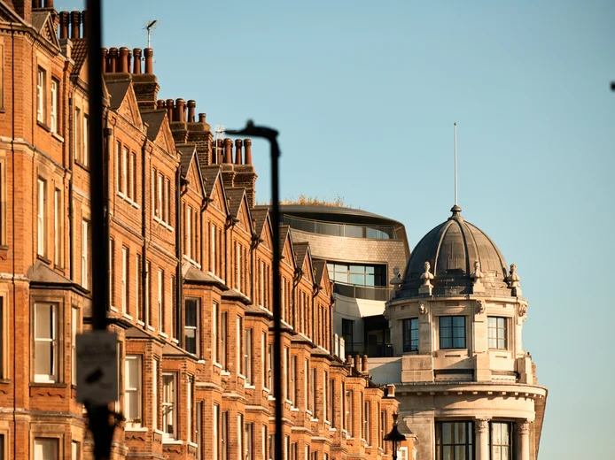 Red brick townhouses with tall chimneys line a quiet street next to a domed heritage building, all beneath an open blue sky.