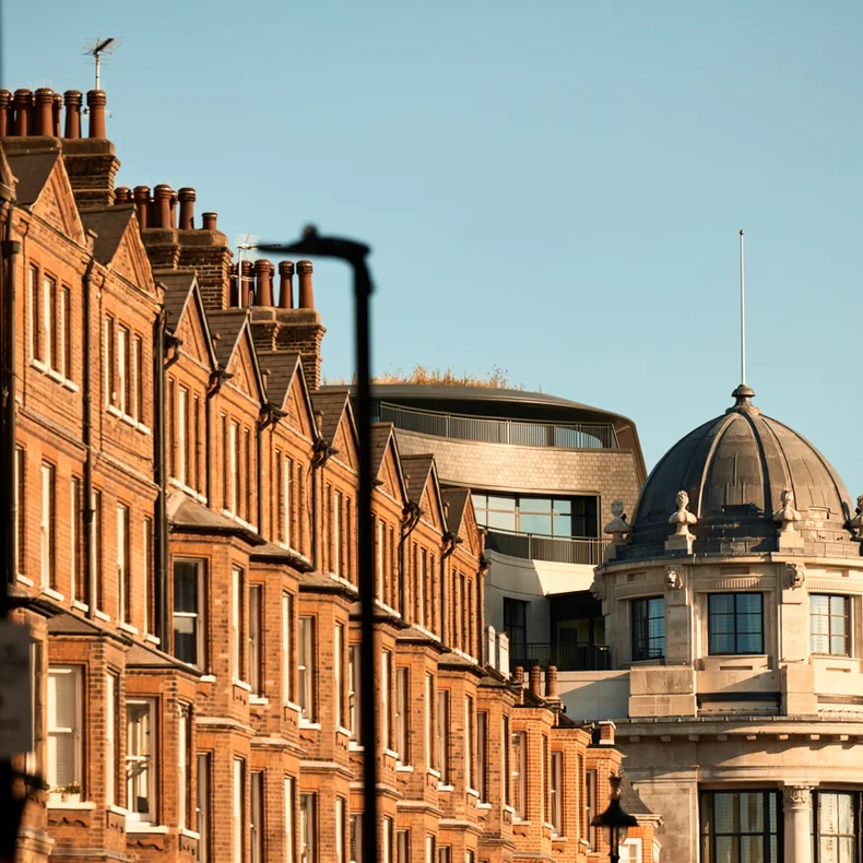 Red brick townhouses with tall chimneys line a quiet street next to a domed heritage building, all beneath an open blue sky.