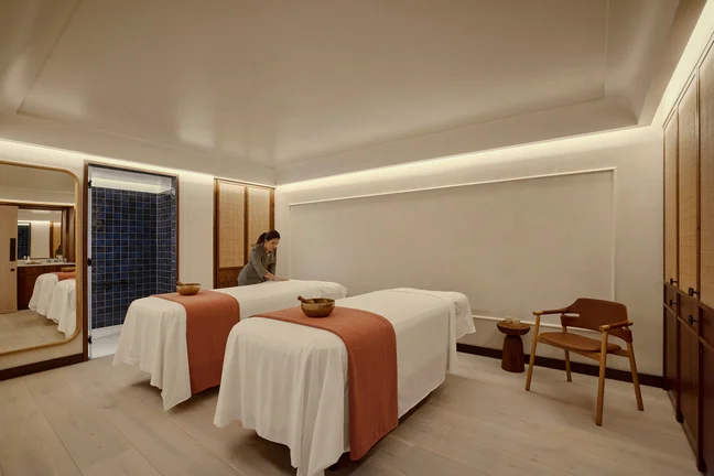 Wellness space featuring two massage tables with white sheets and orange accents, a wooden chair, and a staff member preparing the serene treatment area.