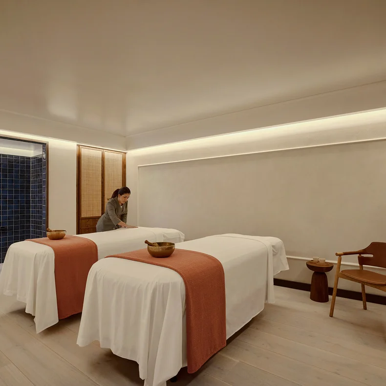 Wellness space featuring two massage tables with white sheets and orange accents, a wooden chair, and a staff member preparing the serene treatment area.