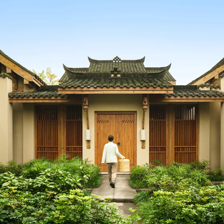 Guest approaches a building with a tiled roof and wooden doors, surrounded by greenery, reflecting traditional architecture and natural landscape.
