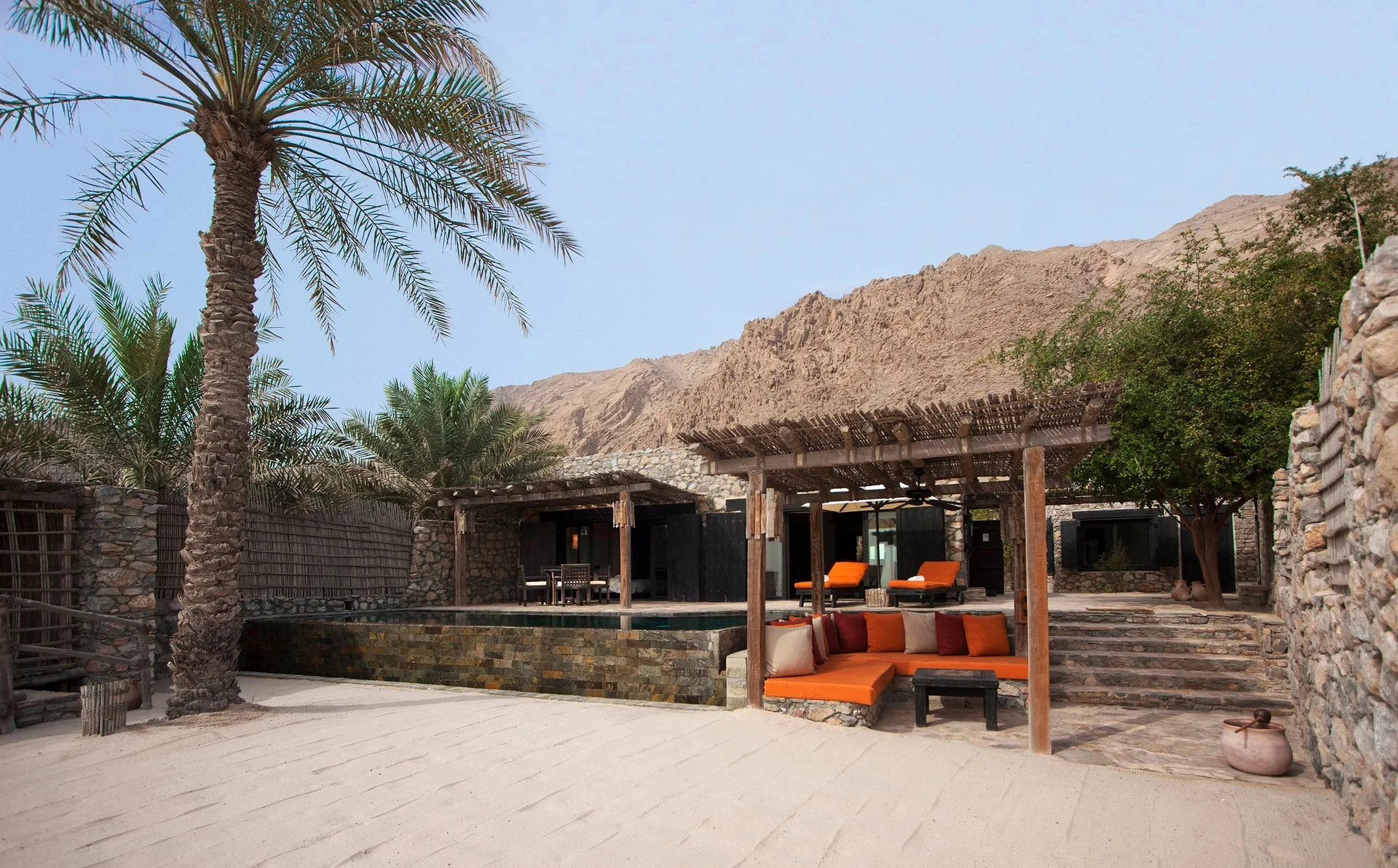 A pool villa suite from its walled sand courtyard, a private pool set into the terrace beside a cane-roofed pergola with orange cushioned seating, boundary walls built from rounded river stone and a date palm above.