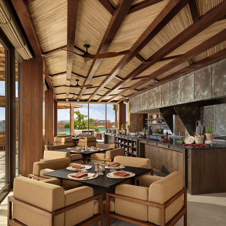 Dining space with wooden tables and chairs, a spacious kitchen island, set tables, and glass doors opening to daylight and greenery beyond.