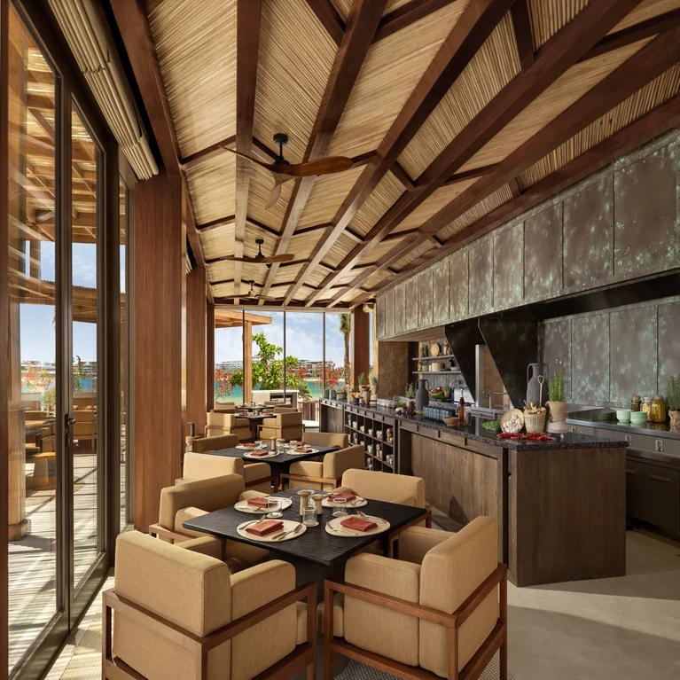 Dining space with wooden tables and chairs, a spacious kitchen island, set tables, and glass doors opening to daylight and greenery beyond.