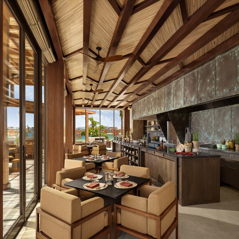 Dining space with wooden tables and chairs, a spacious kitchen island, set tables, and glass doors opening to daylight and greenery beyond.