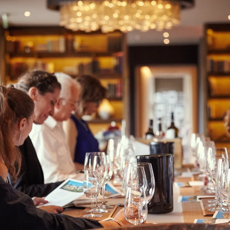 ワイングラス、メニュー、ボトルが並べられたテーブルに客が集まり、心地よく会話できるように設計された柔らかな照明のダイニングスペース。.