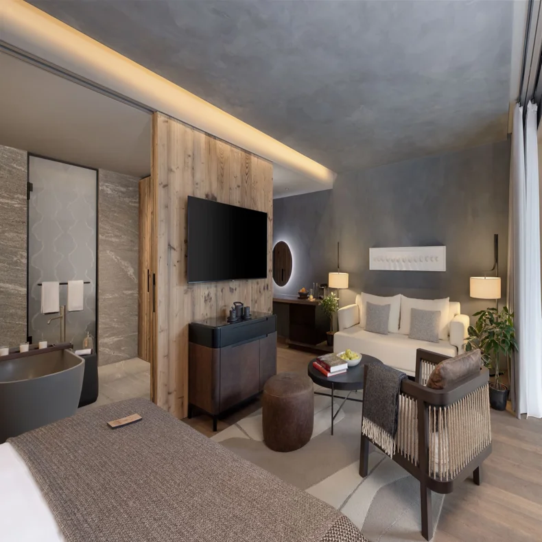 Tranquil room featuring a large bed, comfortable seating, integrated TV, bath, natural wood details and expansive window with sheer curtains that welcome soft daylight.