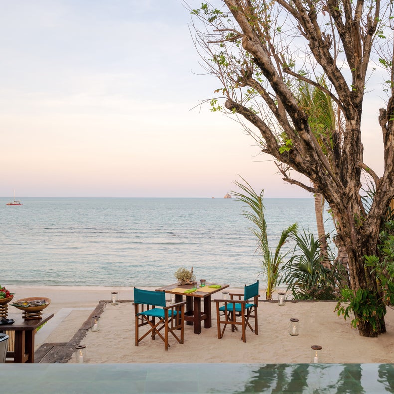 Chef prepares a meal at two tables set on sand, surrounded by trees and greenery, with views of calm sea and soft evening light in the background.