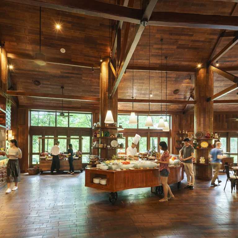 Interior del restaurante con vigas de madera natural, grandes ventanales con luz natural, mesas de bufé con platos frescos y comensales disfrutando de las comidas en un ambiente acogedor.