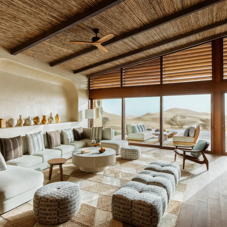 Serene living space featuring floor-to-ceiling windows, earth-toned furnishings, detailed ottomans, artisanal vases, and expansive desert views. Wood and subtle fan for comfort.