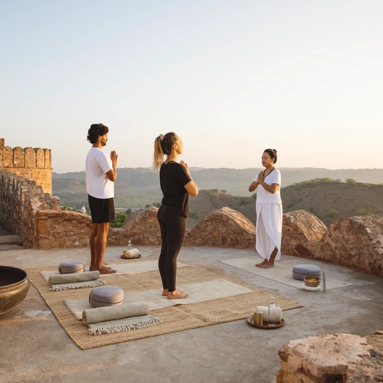 学员们在石头露台上参加晨间瑜伽课程,旁边摆放着冥想坐垫和蜡烛,在柔和的自然光中欣赏山丘和古代遗迹。.