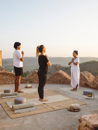 Khách tham gia lớp yoga buổi sáng trên một sân thượng bằng đá, với những chiếc gối thiền và nến gần đó, nhìn ra những ngọn đồi và di tích cổ kính trong ánh sáng tự nhiên dịu nhẹ.