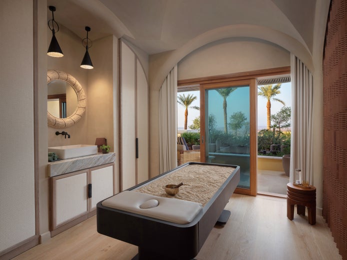 Spa room featuring a massage table, round mirror over a sink, gentle lighting, and glass doors leading to an outdoor patio with palm trees and seating.