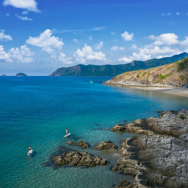 Turquoise ocean meets a rocky shoreline and sandy beach, with two paddleboarders and green hills beneath a blue sky dotted with clouds.