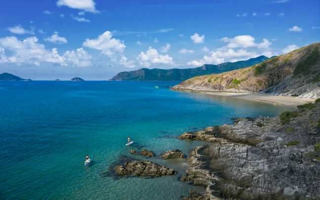 Turquoise ocean meets a rocky shoreline and sandy beach, with two paddleboarders and green hills beneath a blue sky dotted with clouds.