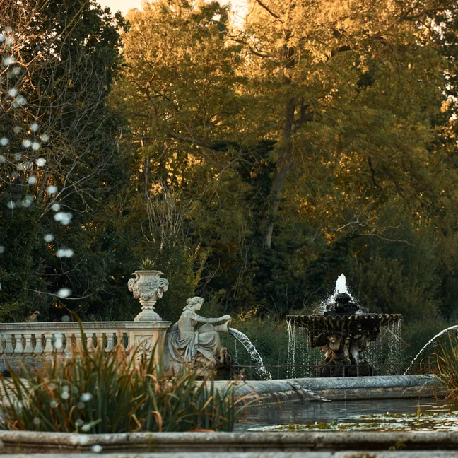 Stone fountain with statues and gentle water jets, encircled by trees and garden greenery in soft sunset light.