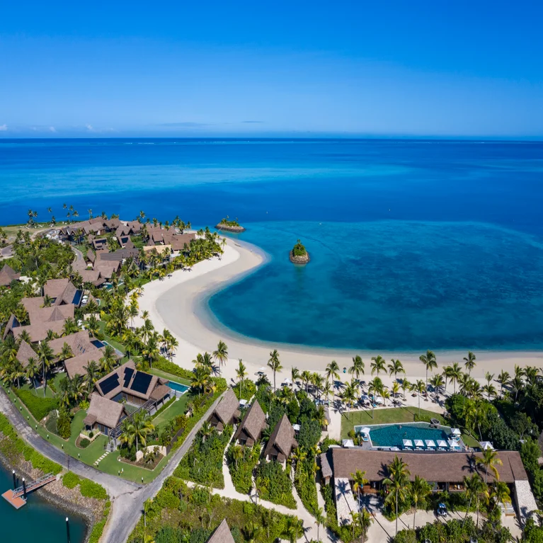 Thatched-roof villas line a white sand beach with palm trees and a swimming pool overlooking a blue lagoon.