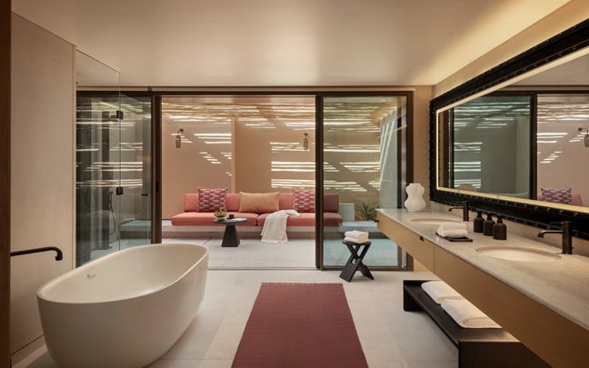 Peaceful bath area featuring a freestanding tub, dual sinks, and mirror; glass doors open to a patio with pastel seating and soft lighting. Bright natural daylight fills the space.