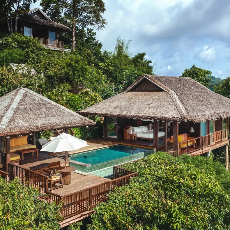Villa with sustainable design on a hillside, featuring an infinity pool and sun loungers, surrounded by tropical plants beneath a partly cloudy sky.