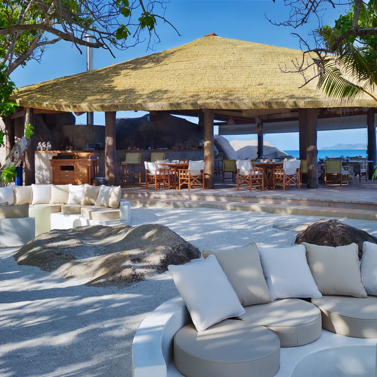 树下沙地上的环形软垫座椅,毗邻茅草屋顶的开放式凉亭,可欣赏海景,设计舒适、放松。.