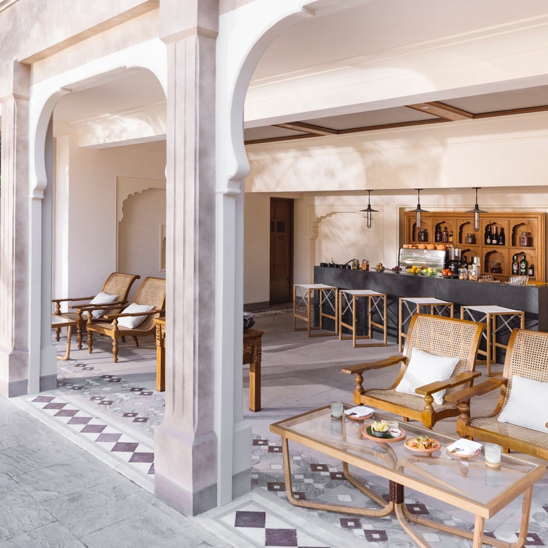 Patio al aire libre con columnas arqueadas, sillas de madera, mesa de café de cristal que ofrece aperitivos, y un mostrador de bar con taburetes y estanterías de botellas al fondo.
