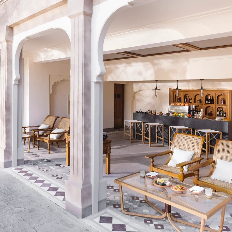 Open-air patio with arched columns, wooden chairs, glass coffee table offering snacks, and a bar counter with stools and shelves of bottles in the background.
