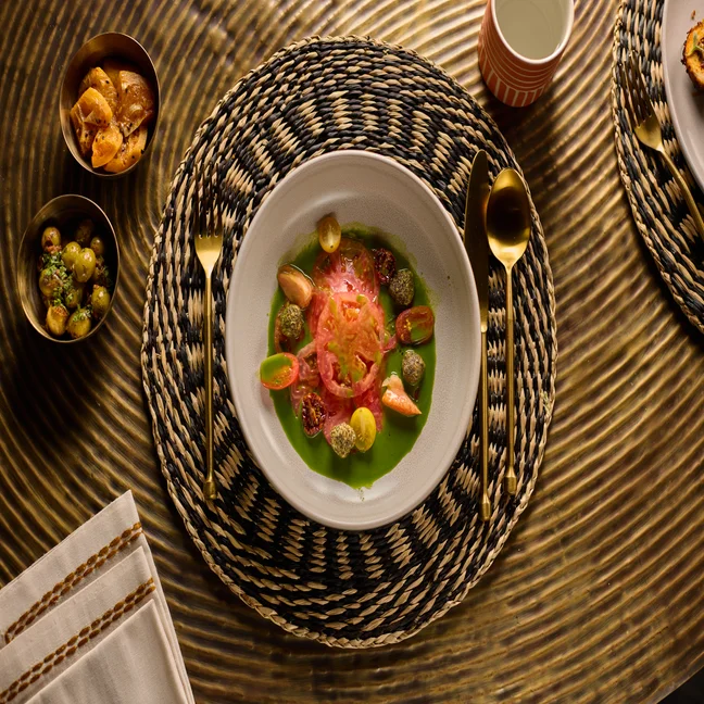 Seasonal vegetables with tomato sauce are served on a woven placemat. Gold cutlery, olives, a cup, and a bowl of chips rest on a golden textured table.