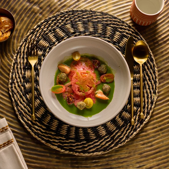Seasonal vegetables with tomato sauce are served on a woven placemat. Gold cutlery, olives, a cup, and a bowl of chips rest on a golden textured table.