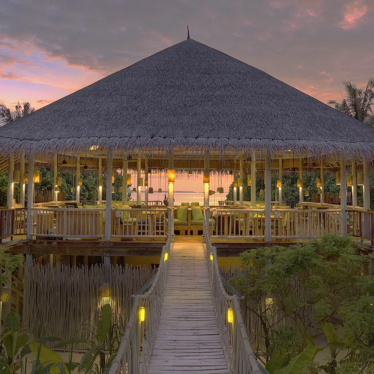 El comedor, de planta abierta, se asienta sobre una vegetación vibrante y se ilumina con una suave luz al atardecer, creando un ambiente sereno en armonía con la naturaleza.