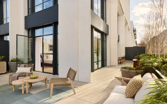 Patio with natural wood seating and tables on a tiled terrace, adjacent to glass doors. Green plants enhance the space, inviting connection with nature and daylight.